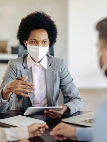 Bank manager with face mask using touchpad while talking to her clients on a meeting in the office.