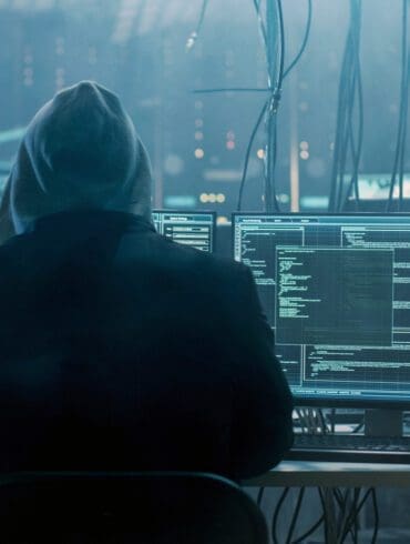 Dangerous Hooded Hacker Breaks into Government Data Servers and Infects Their System with a Virus. His Hideout Place has Dark Atmosphere, Multiple Displays, Cables Everywhere.