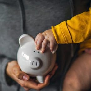 A baby holds on to the ear of a piggy bank while being held by its mother.