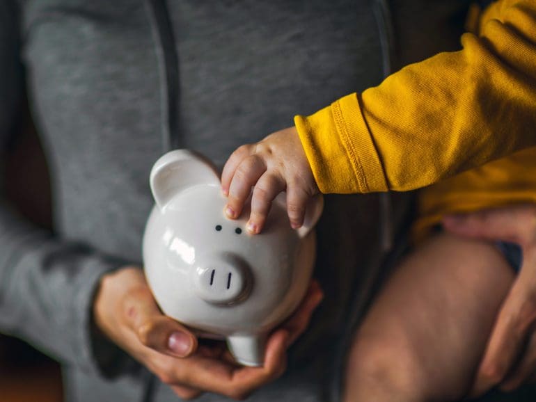 A baby holds on to the ear of a piggy bank while being held by its mother.