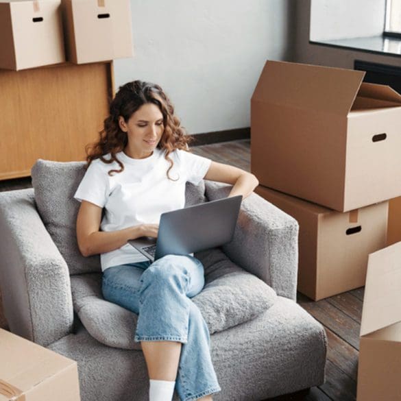 Happy young hispanic woman renting apartment online. Girl among cardboard boxes is using laptop and smiling. Happy homeowner is sitting in armchair with pc. Woman about to move.