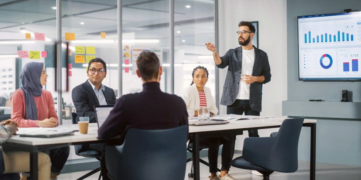 Diverse Modern Office: Businessman Leads Business Meeting with Managers, Talks, uses Presentation TV with Statistics, Infographics. Digital Entrepreneurs Work on e-Commerce Project.