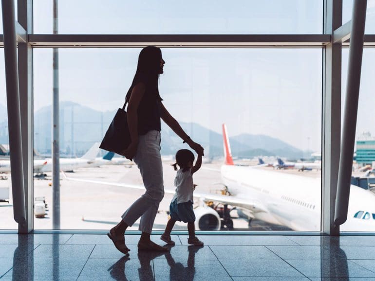 Silhouette of joyful young Asian mother holding hands of cute little daughter looking at airplane through window at the airport while waiting for departure