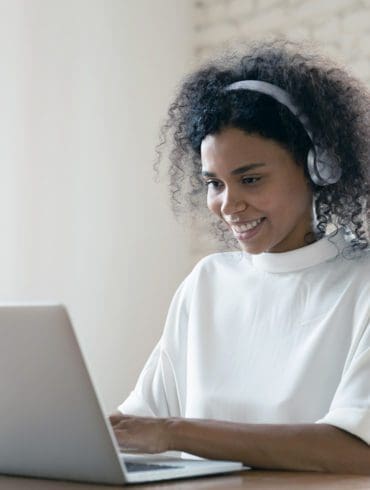 Smiling African American woman wearing headset using laptop in modern office, looking at screen and typing, listening to music, interpreter working online, learning computer course, lecture