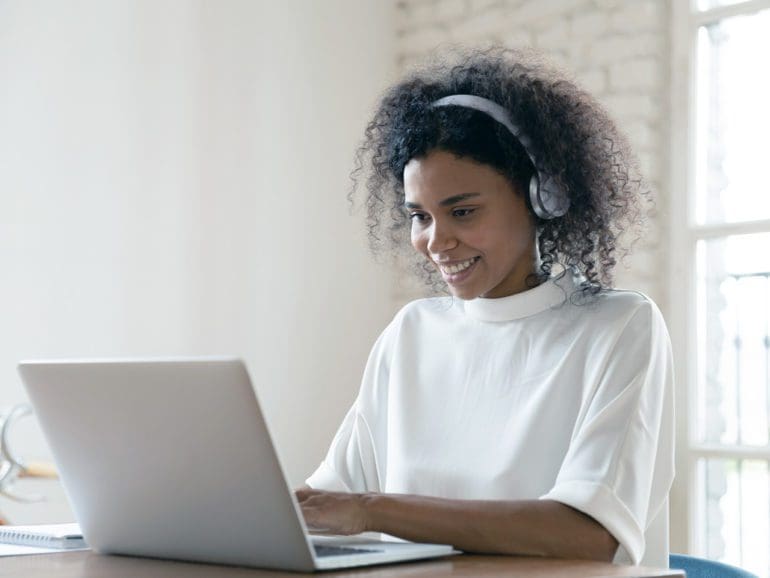 Smiling African American woman wearing headset using laptop in modern office, looking at screen and typing, listening to music, interpreter working online, learning computer course, lecture