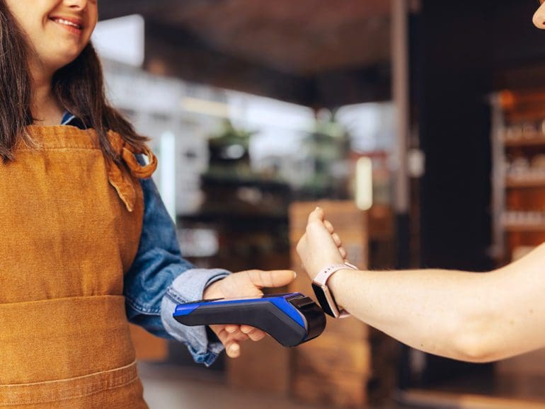 Happy customer scanning her smartwatch on a credit card machine to pay her bill in a cafe. Cheerful woman doing a cashless and contactless transaction using NFC technology.