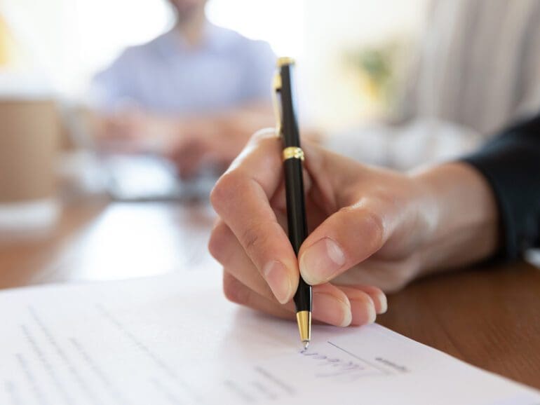 Close up young woman putting signature on banking loan document in office. Successful businesswoman signing contract or purchase document with partners at meeting after negotiations and making edits.