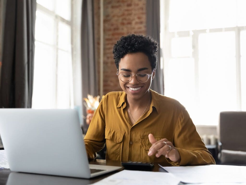 Happy young Afro American entrepreneur woman in glasses counting profit, on calculator at laptop computer, analyzing benefits, enjoying financial success, job high result, smiling