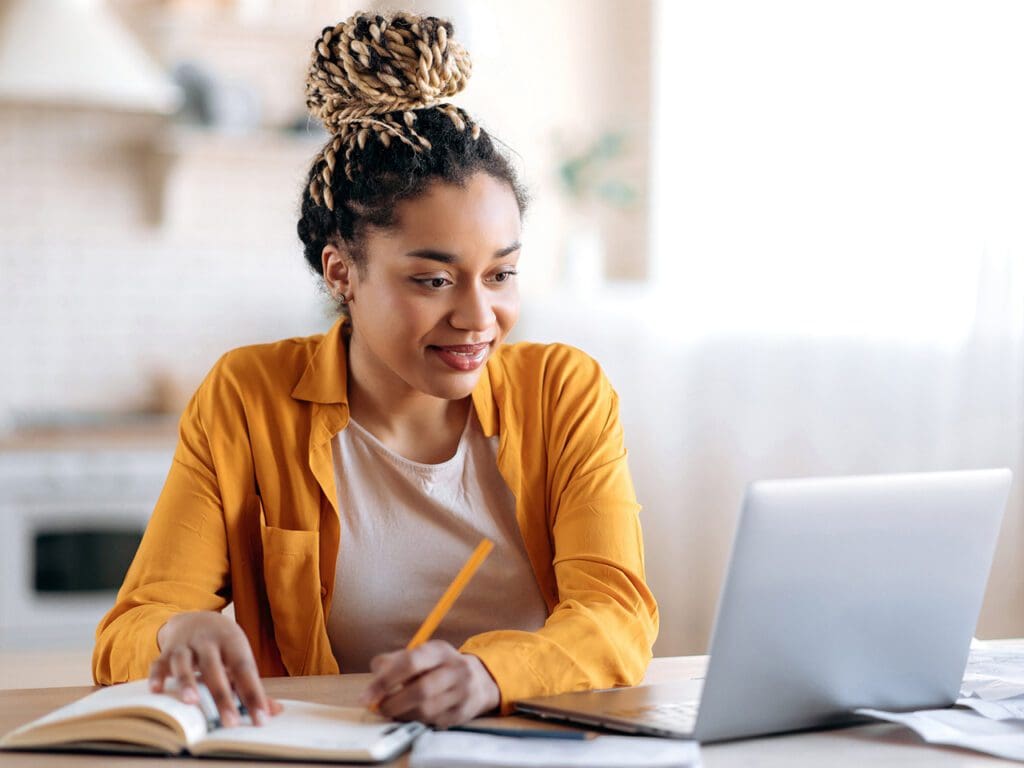 Focused cute stylish african american female student with afro dreadlocks, studying remotely from home, using a laptop, taking notes on notepad during online lesson, e-learning concept, smiling