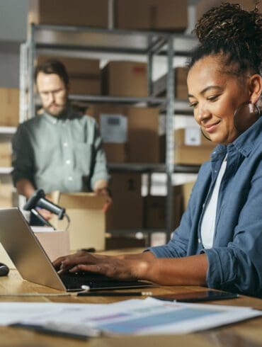 Diverse Male and Female Warehouse Inventory Managers Talking, Using Laptop Computer and Checking Retail Stock. Rows of Shelves Full of Cardboard Box Packages in the Background.